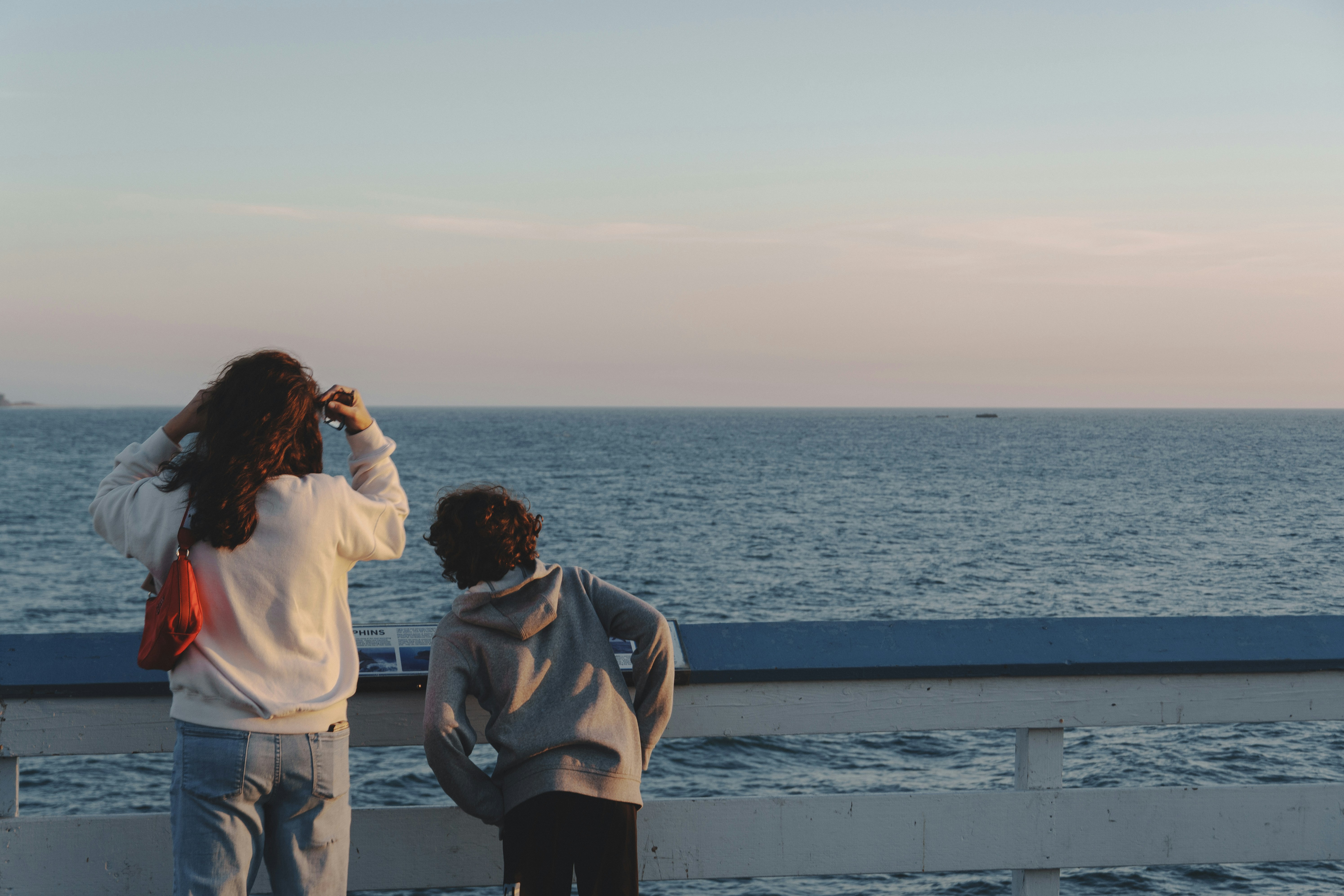 Two people, one adult and one child, stand by a wooden railing overlooking a calm ocean at sunset, gazing toward the horizon